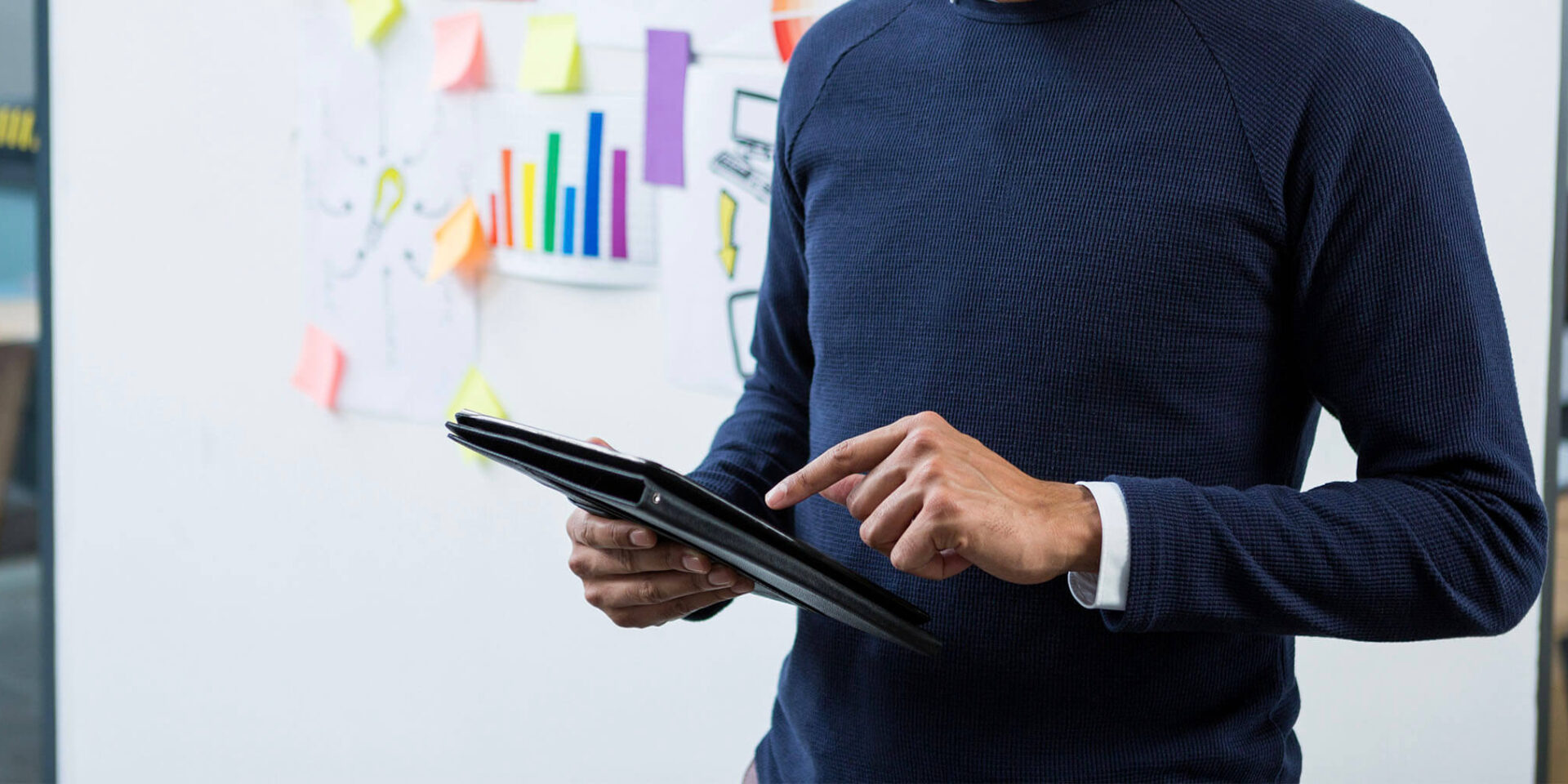 A business professional standing with a tablet, engaged in a presentation with colorful charts and notes in the background.