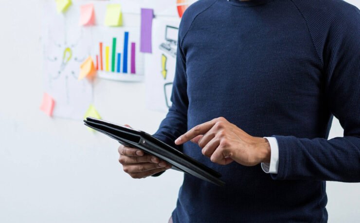 A business professional standing with a tablet, engaged in a presentation with colorful charts and notes in the background.