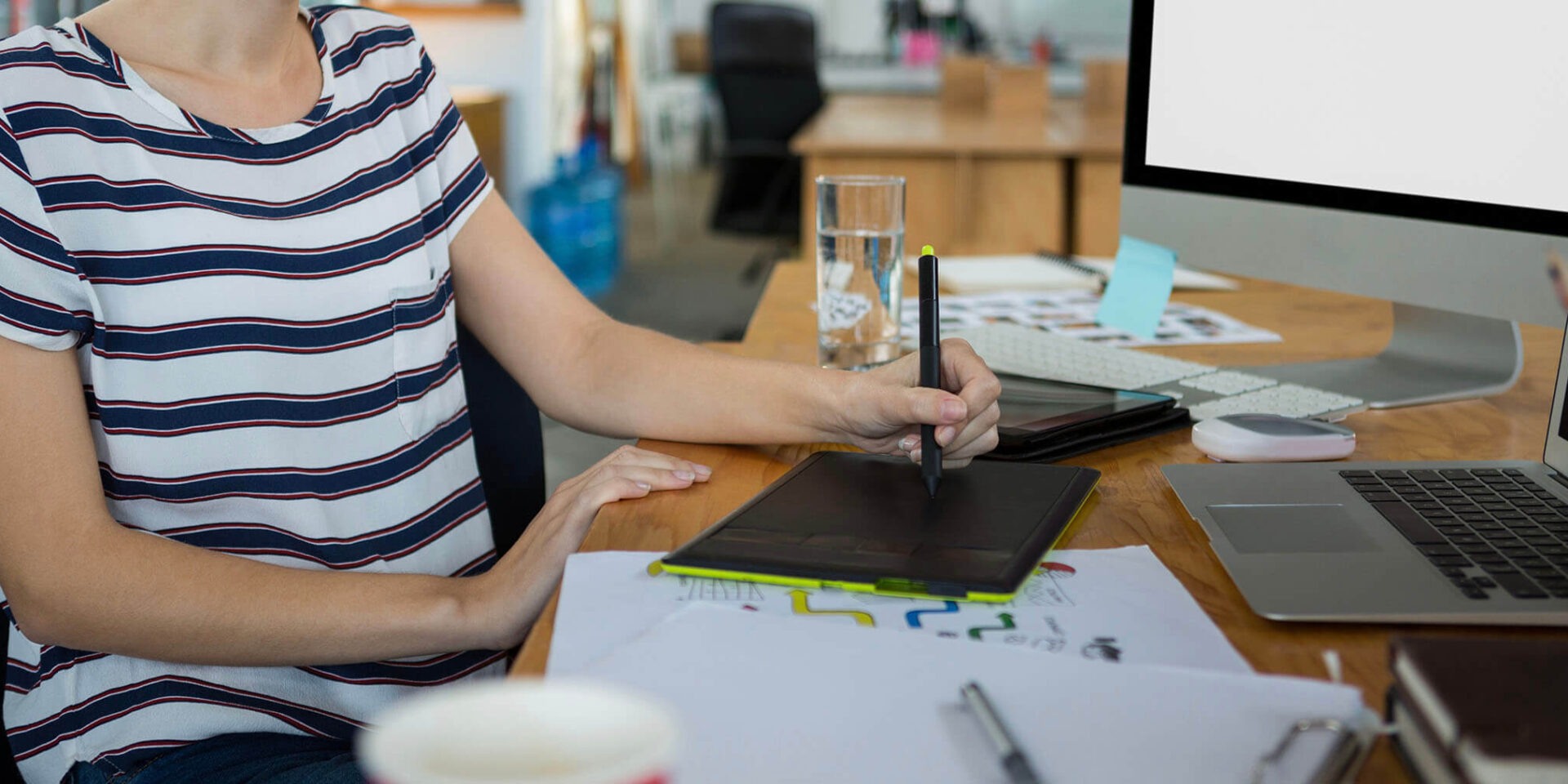 An individual in a striped shirt using a digital tablet at a desk with a laptop and papers scattered on the table.