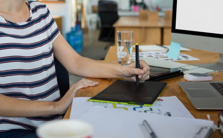 An individual in a striped shirt using a digital tablet at a desk with a laptop and papers scattered on the table.