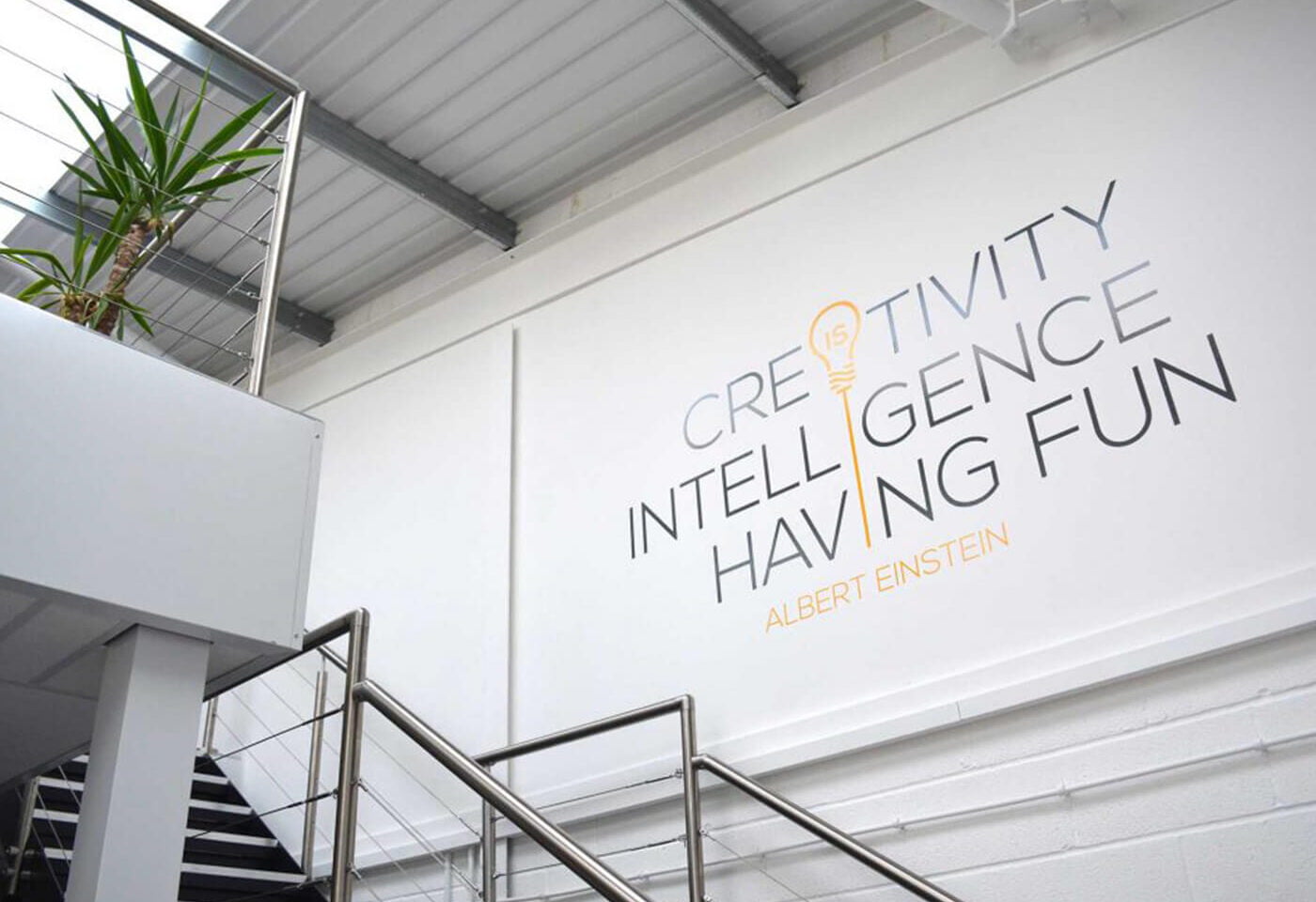A modern office interior featuring a wall with the words 'Creativity, Intelligence, Having Fun' attributed to Albert Einstein, surrounded by a decorative plant and staircase.