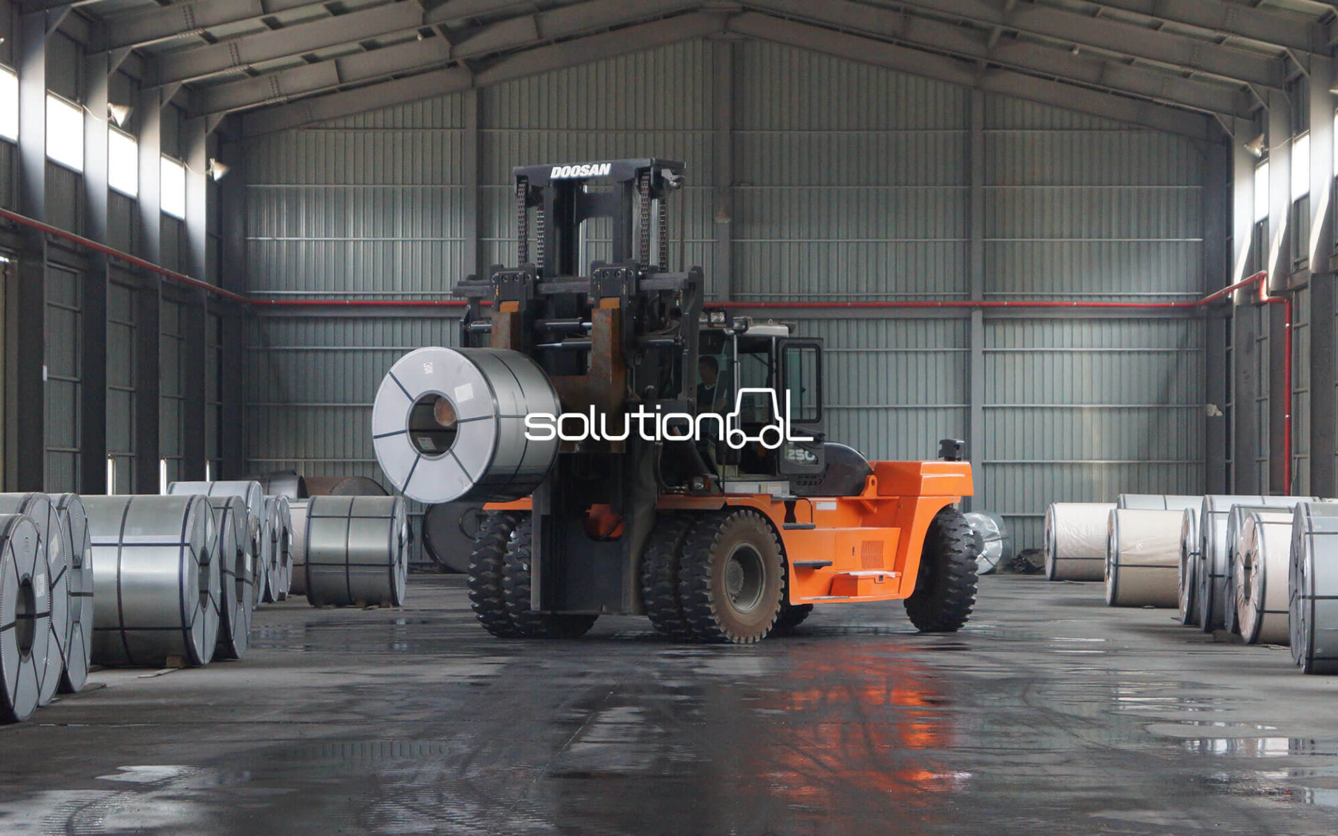 A heavy-duty forklift moving metal coils inside a spacious industrial warehouse.