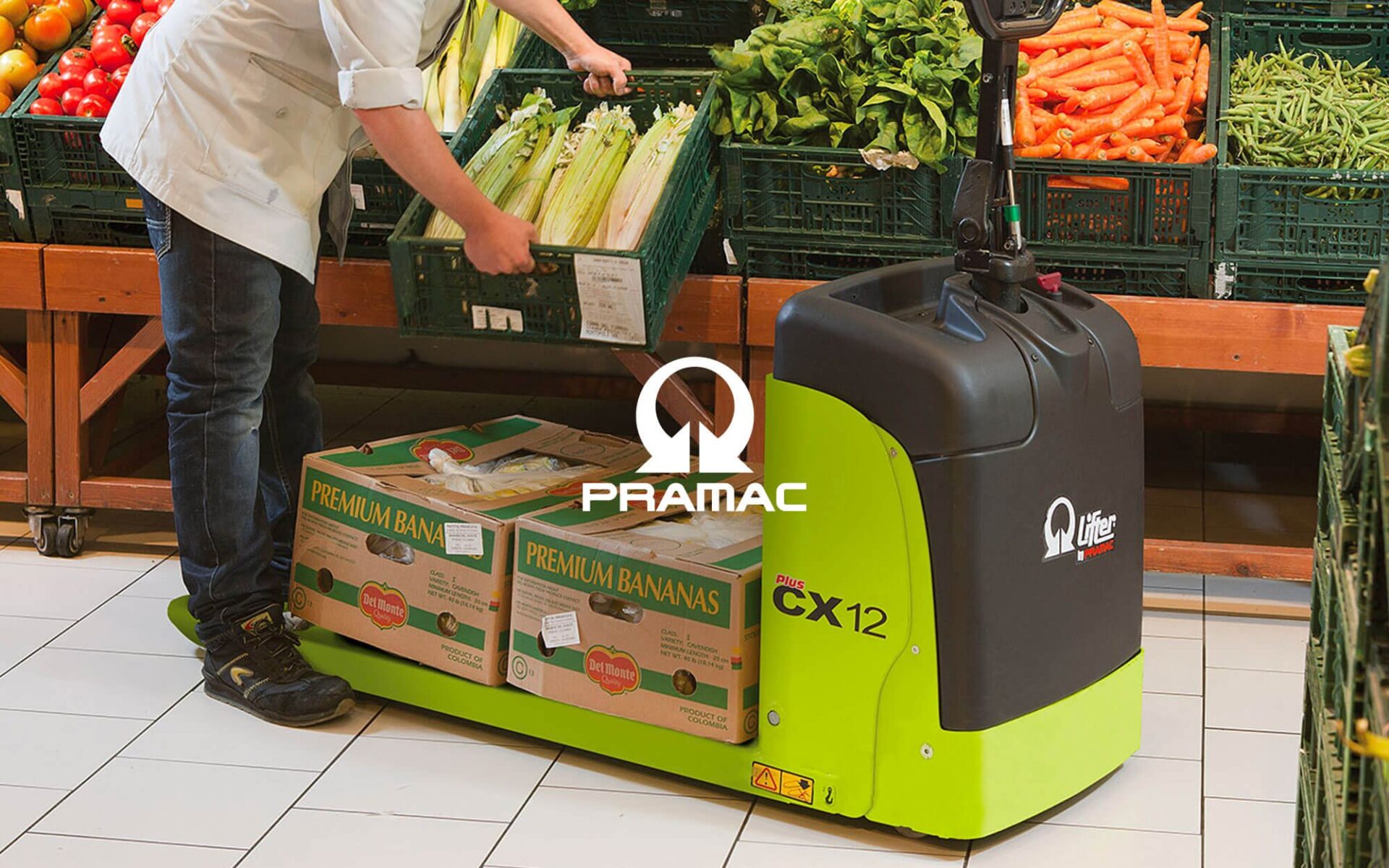 A worker using a Pramac CX12 electric pallet truck to transport boxes in a grocery store filled with fresh produce.