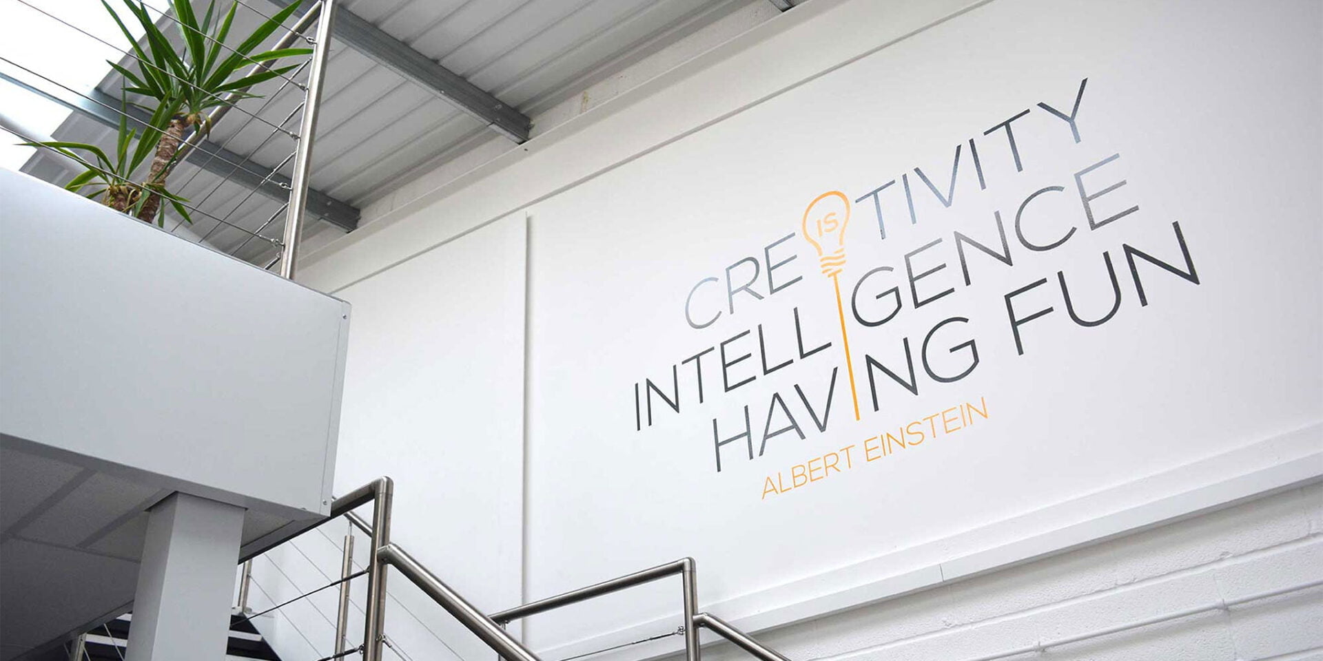 A bright office interior featuring a wall with the words 'Creativity', 'Intelligence', and 'Having Fun' in stylish typography, alongside a staircase and indoor plants.