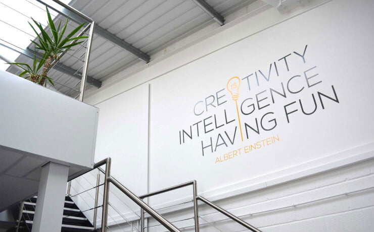 A bright office interior featuring a wall with the words 'Creativity', 'Intelligence', and 'Having Fun' in stylish typography, alongside a staircase and indoor plants.