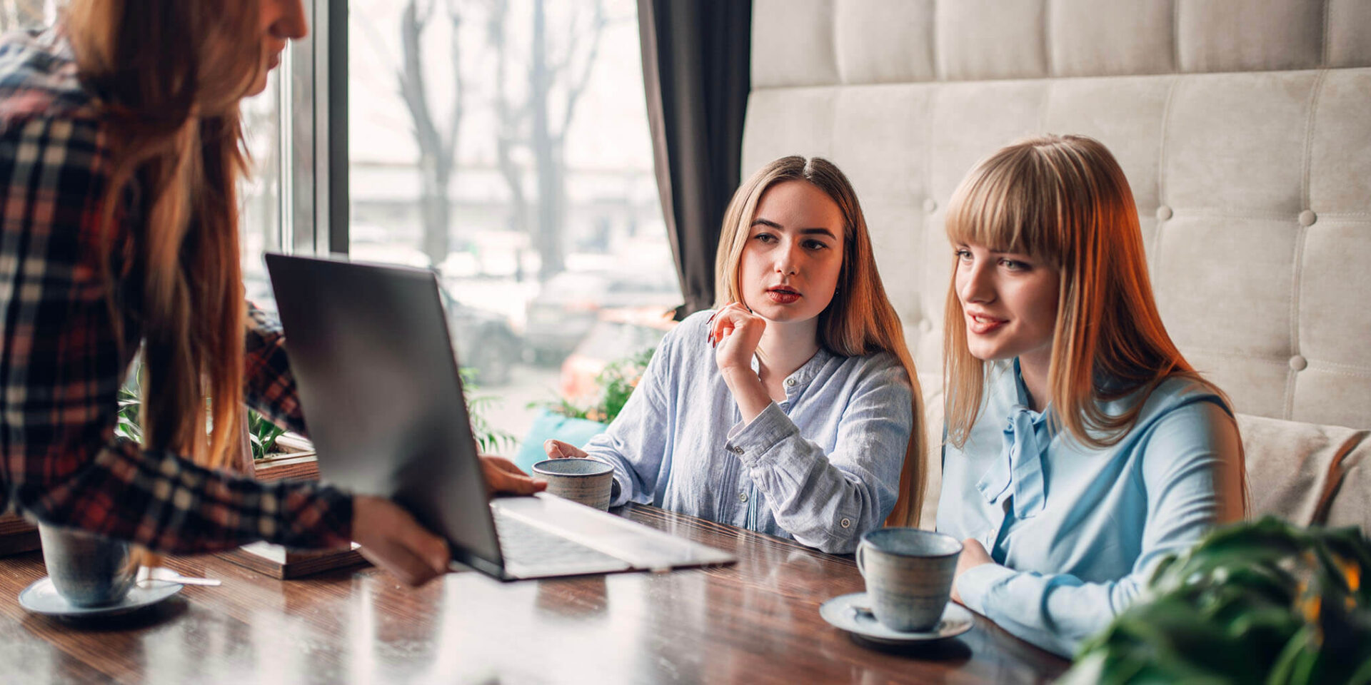 Two women attentively listening and discussing while a third person shows them something on a laptop in a warm café setting.