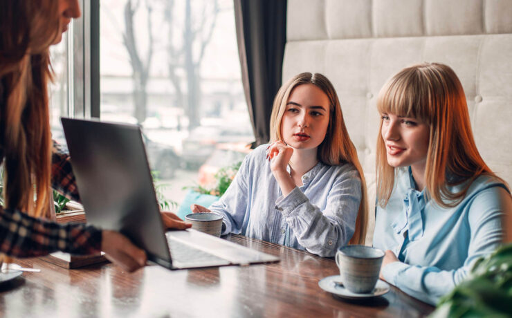 Two women attentively listening and discussing while a third person shows them something on a laptop in a warm café setting.