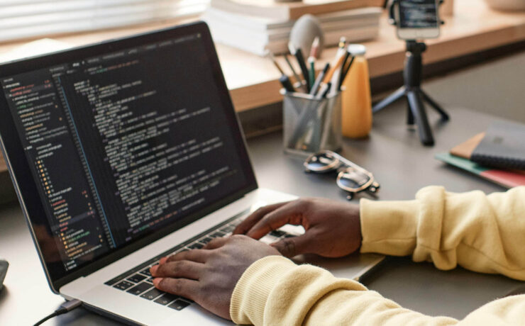 Close-up of hands typing on a laptop with code on the screen and a desk with office supplies in the background.