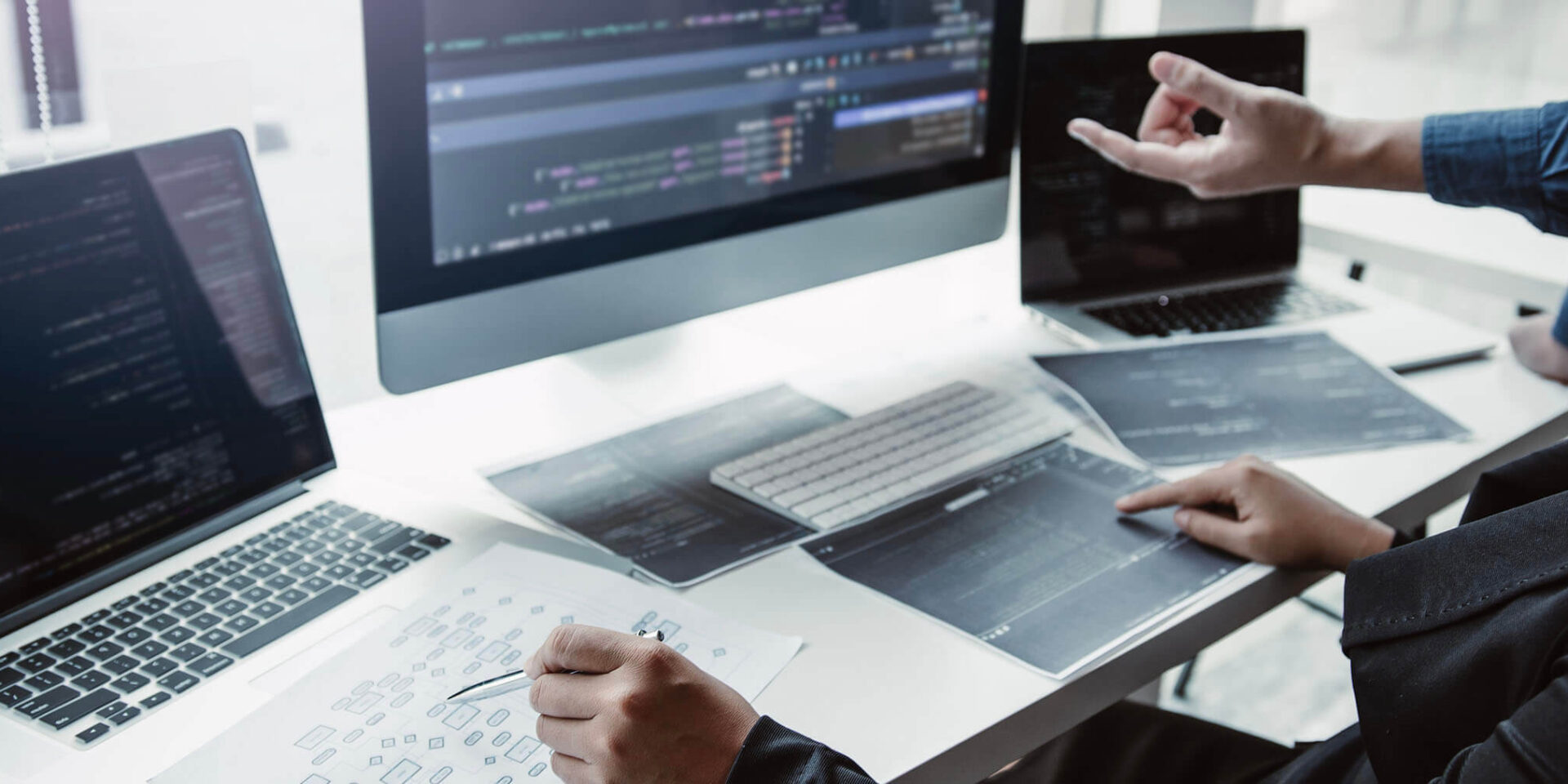 A close-up of a modern coding workstation featuring multiple screens displaying code, with a person discussing and pointing at the screen.
