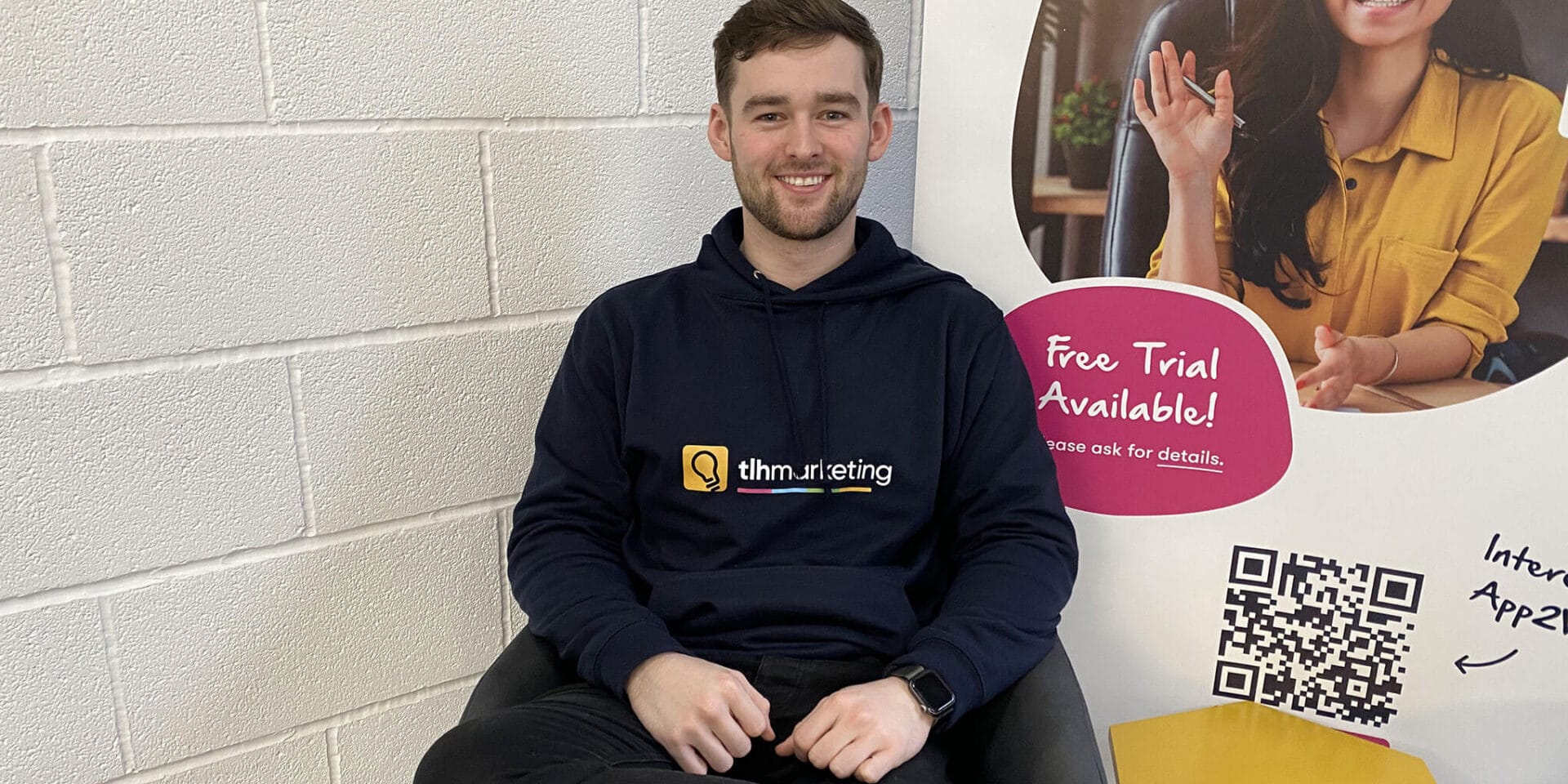 A young man wearing a hoodie sits comfortably in a chair in a modern workspace, with a promotional sign indicating a free trial available.